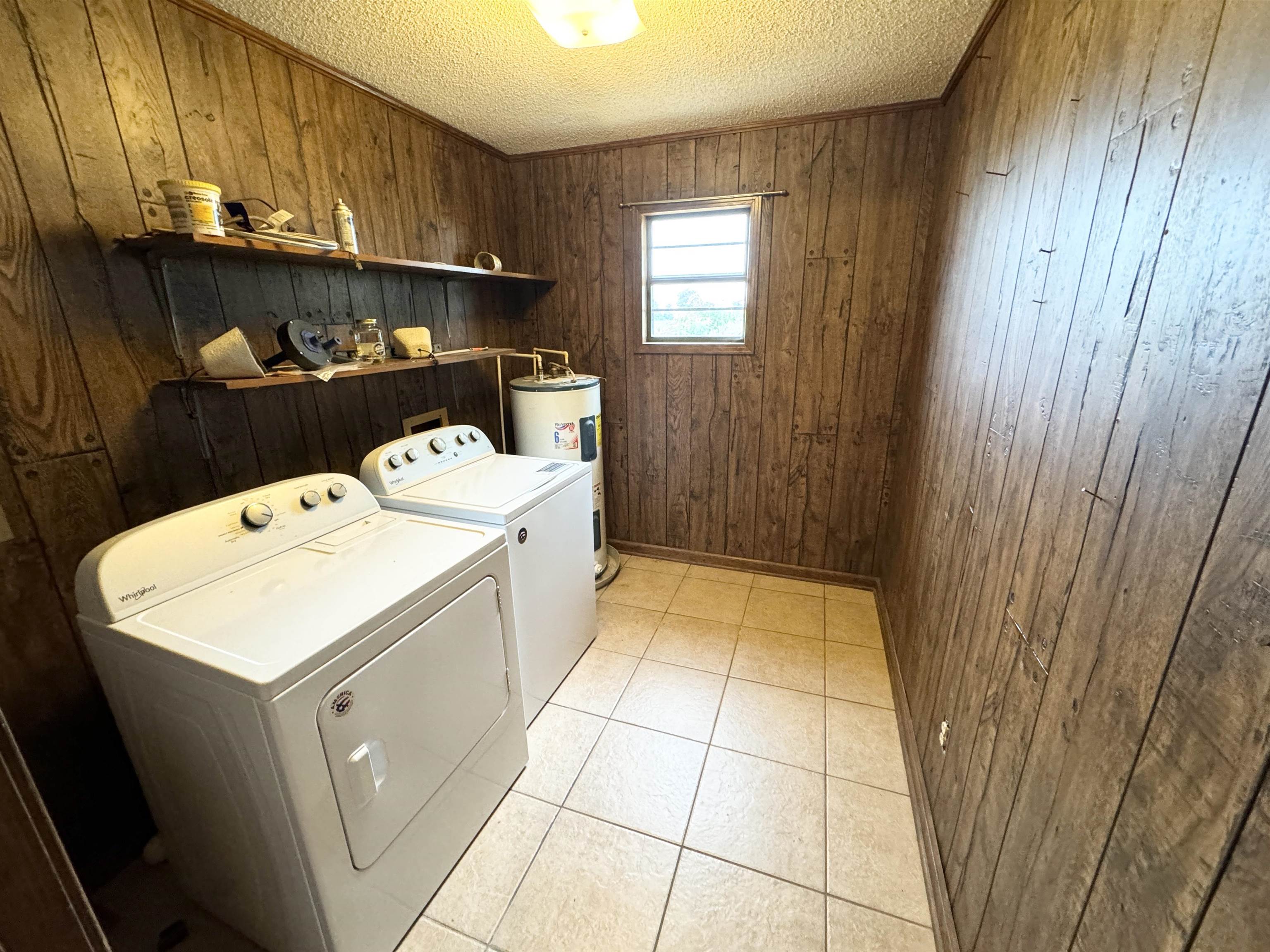 Laundry room with wooden walls, electric water heater, washer and clothes dryer, light tile patterned floors, and a textured ceiling