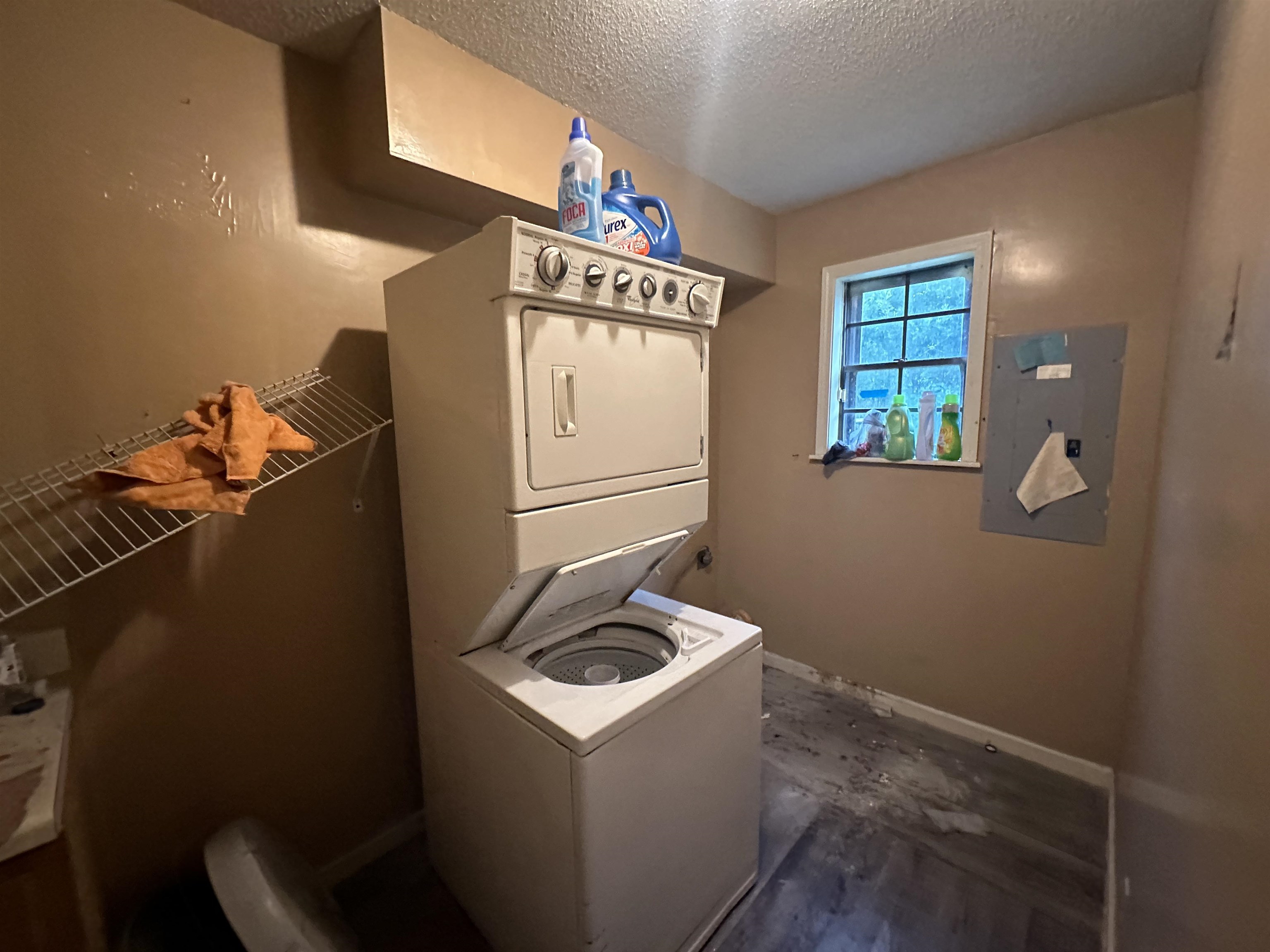 Laundry area with a textured ceiling, stacked washer and clothes dryer, and electric panel