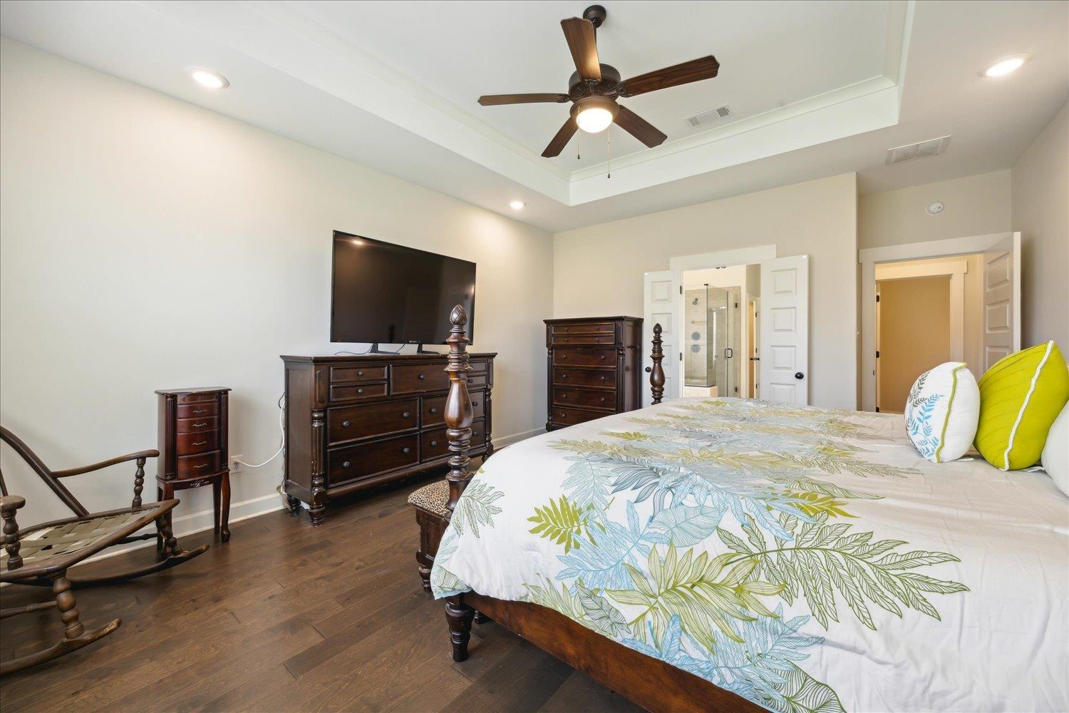 Bedroom with dark wood-type flooring, ceiling fan, ensuite bath, and a tray ceiling