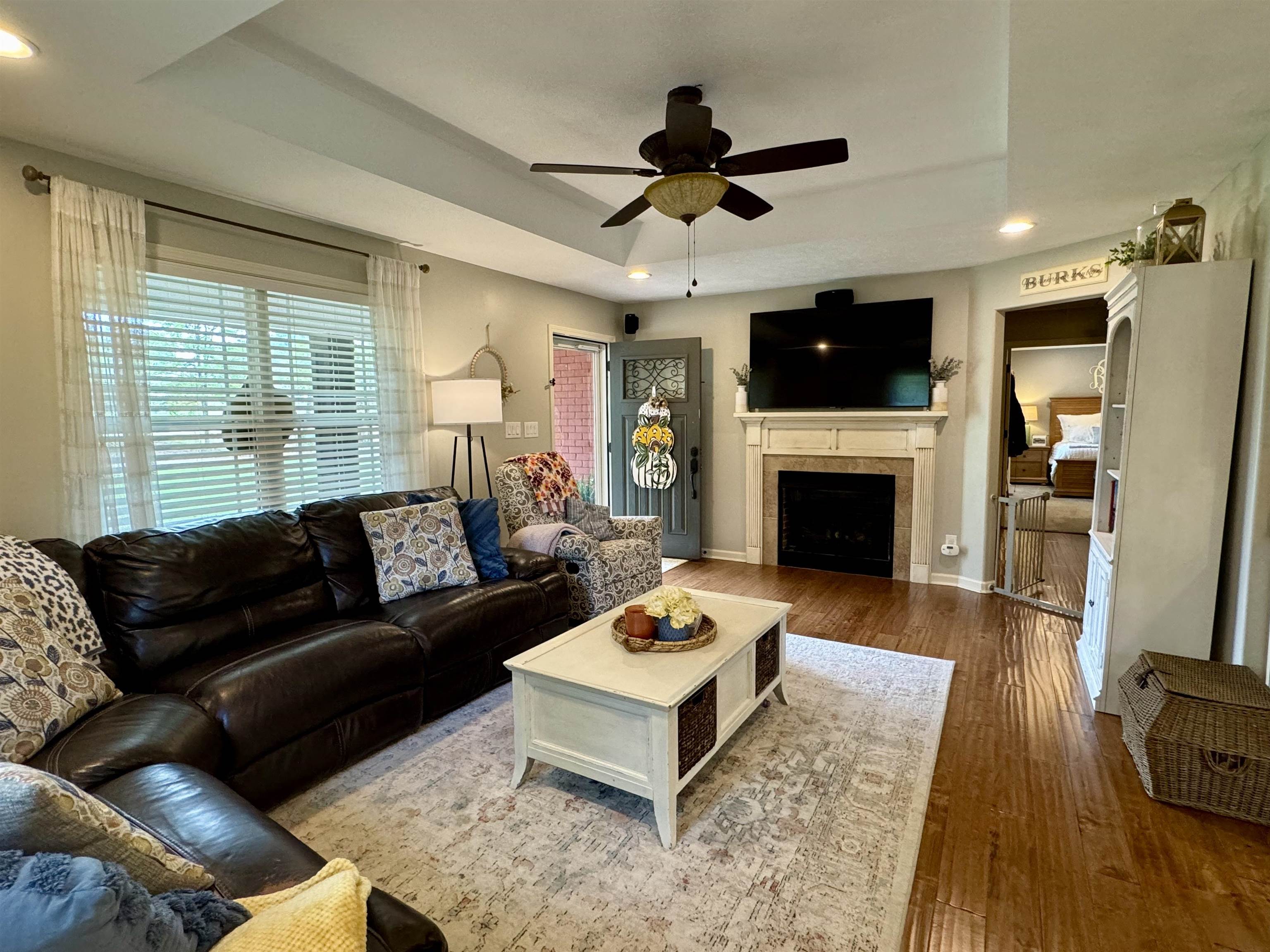 Living room with hardwood / wood-style floors, a tray ceiling, a tiled fireplace, and ceiling fan