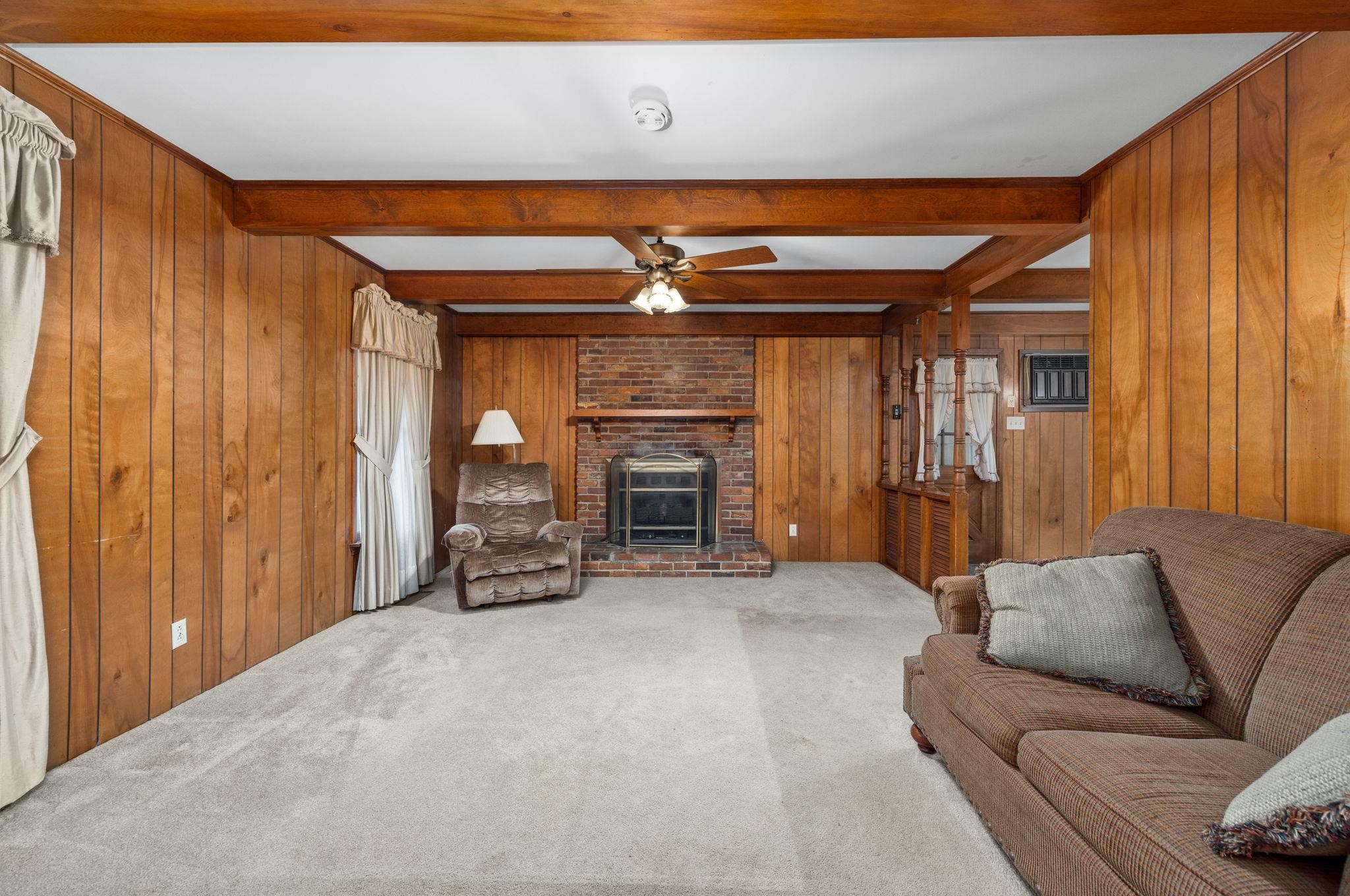 Living room featuring beam ceiling, ceiling fan, carpet floors, wooden walls, and a fireplace