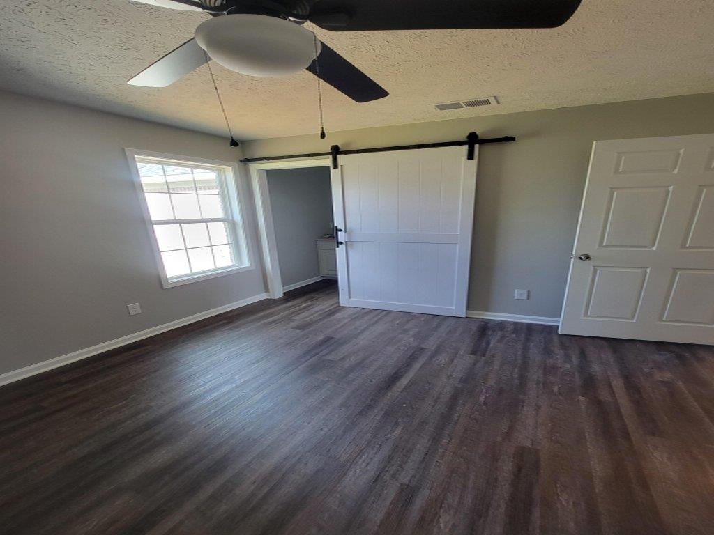 Unfurnished bedroom featuring a textured ceiling, a barn door, dark hardwood / wood-style floors, and ceiling fan