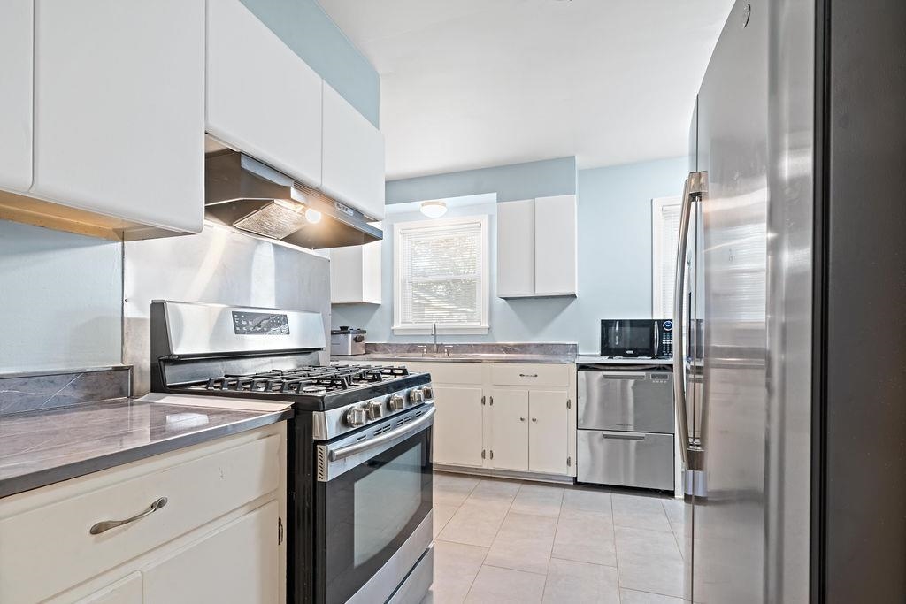 Kitchen with ventilation hood, stainless steel appliances, sink, white cabinets, and light tile patterned flooring