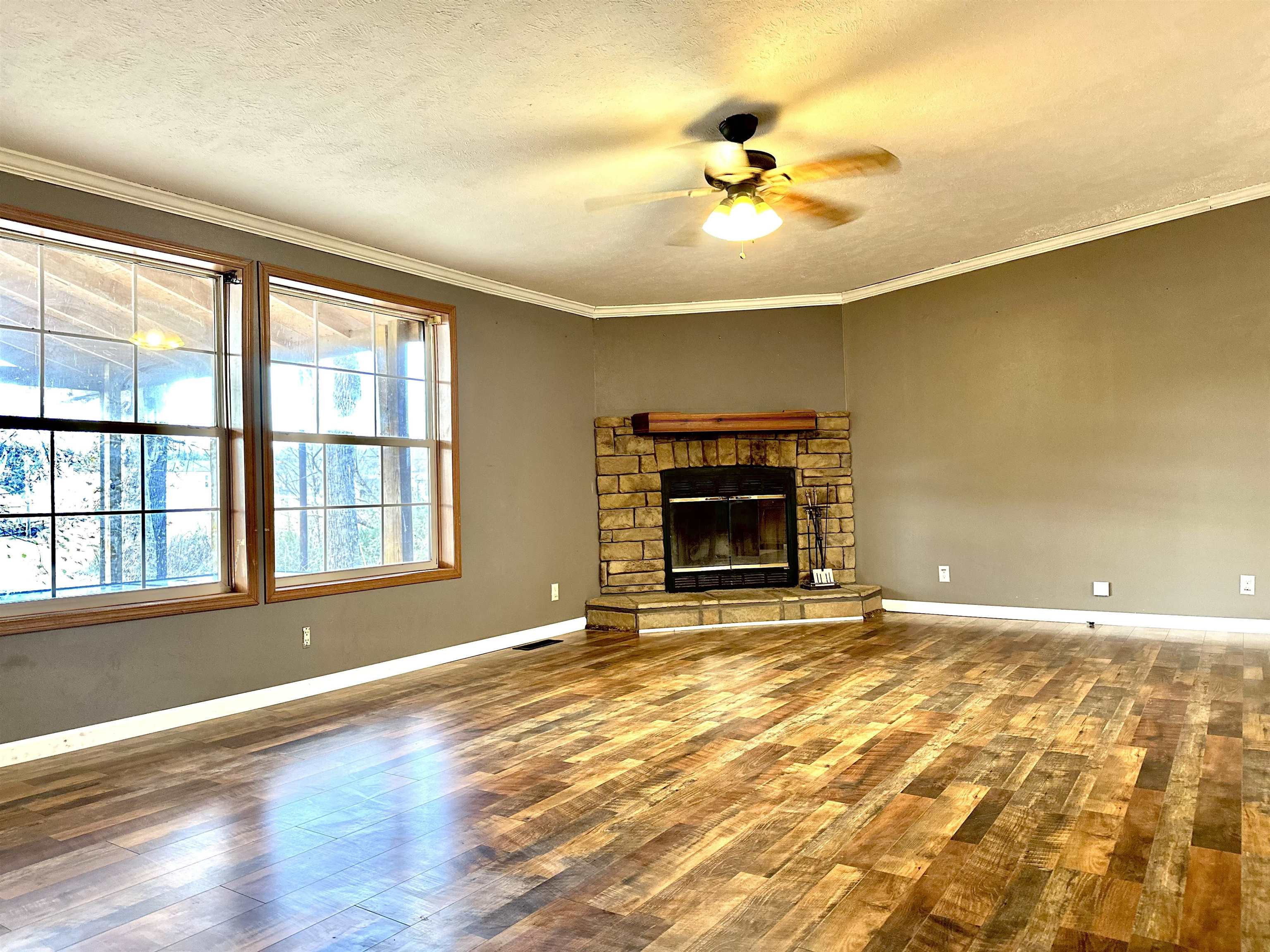 Unfurnished living room with wood-type flooring, a stone fireplace, ceiling fan, and crown molding