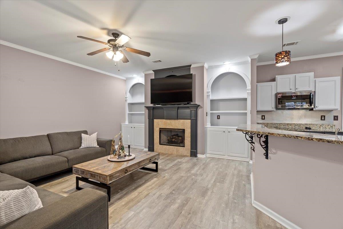 Living room with built in shelves, ceiling fan, crown molding, light hardwood / wood-style floors, and a tiled fireplace