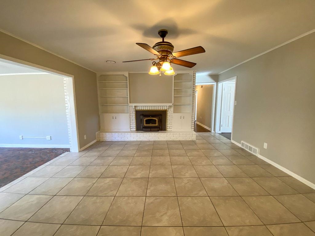 Unfurnished living room with built in shelves, ceiling fan, ornamental molding, and a brick fireplace